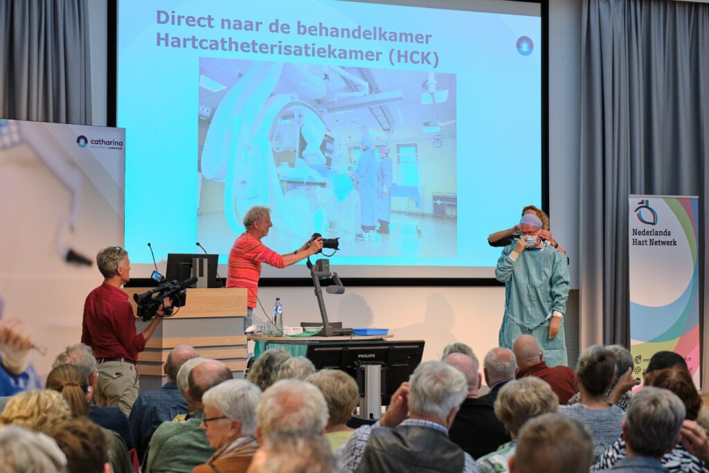 Presentator in medische kleding spreekt op podium, wordt gefilmd; publiek kijkt naar projectie over hartinfarct in het Nederlands.