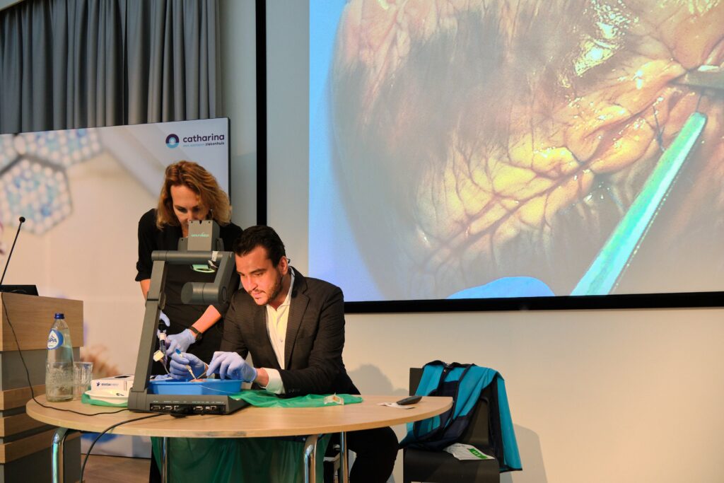 Man demonstreert medische handeling op model aan tafel met vrouwelijke assistent; op scherm hersenoperatie geprojecteerd.