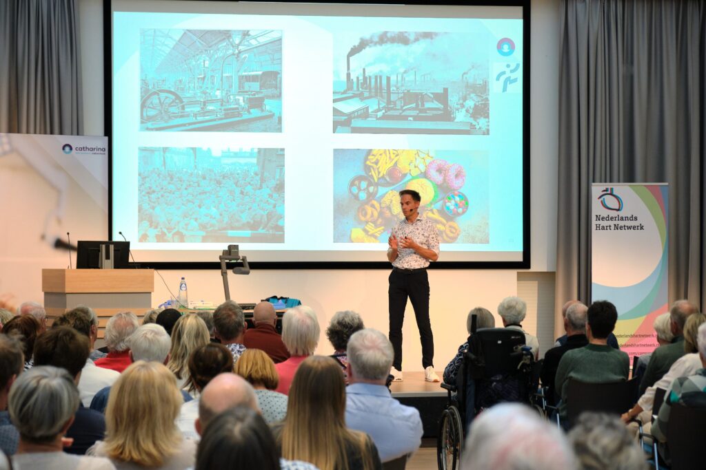 Een man geeft een publiekslezing op het podium. Achter hem een scherm met historische beelden. Publiek en banners aanwezig.