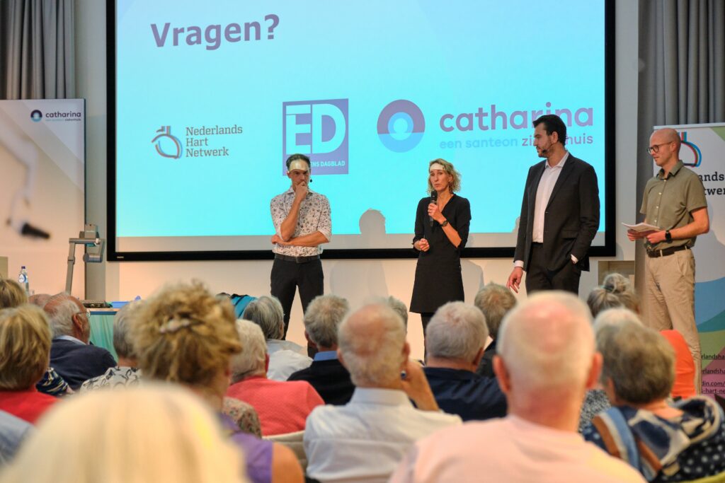 Vier mensen staan op een podium voor publiek bij een publiekslezing. Op het scherm achter hen staat "Vragen?" met logo's.