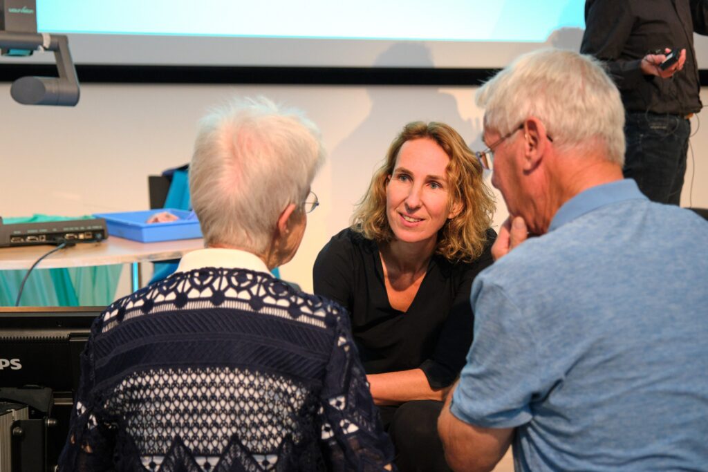 Vrouw met krullen praat met twee oudere volwassenen in een lichte zaal tijdens publiekslezing over hartinfarct; beamer en scherm.
