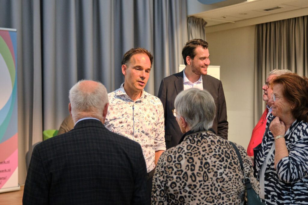 Groep volwassenen praat binnen bij een podium met grijze gordijnen en kleurrijk spandoek tijdens publiekslezing over hartinfarct.