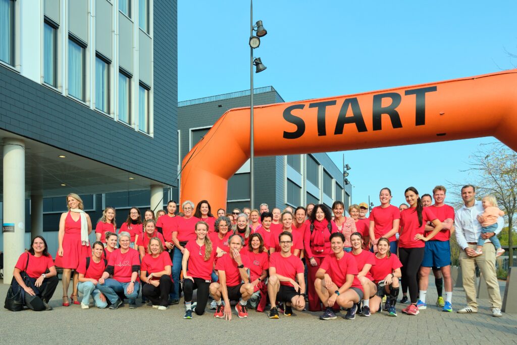 Groep vrouwen in rode shirts poseert voor oranje START-boog bij modern gebouw, klaar voor run tegen hartklachten.