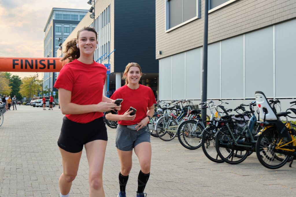 Twee vrouwen in rode sportkleding rennen met telefoons richting een finishlijn op een stadsstraat met geparkeerde fietsen.