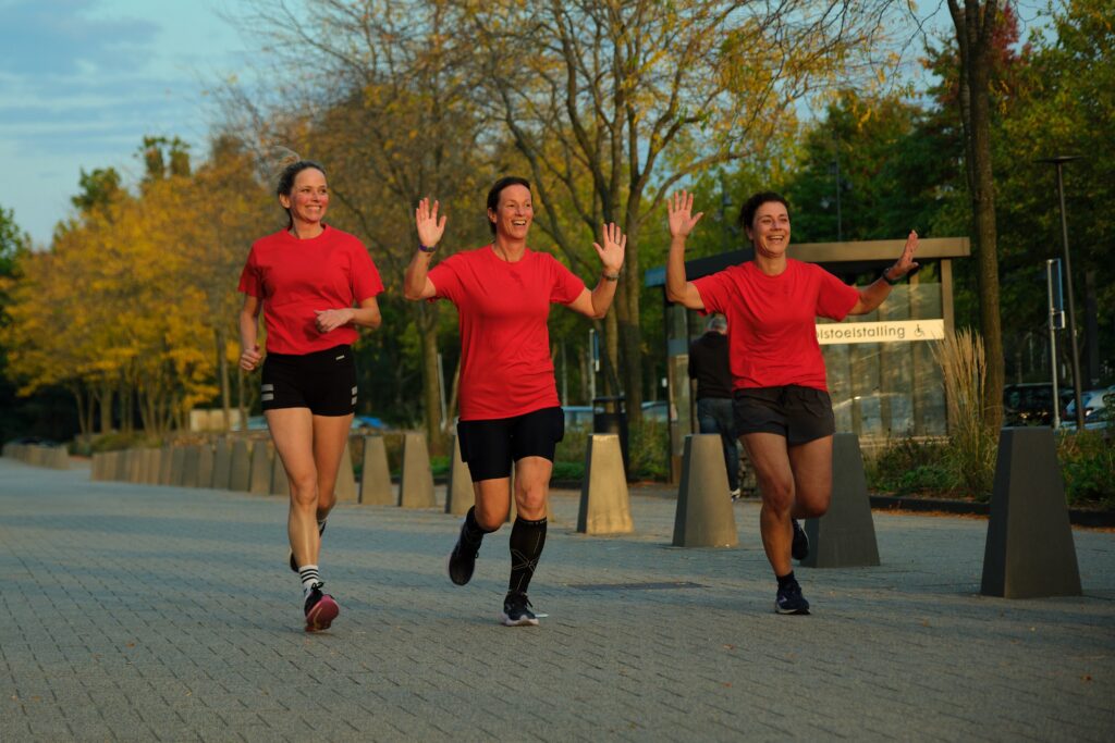 Drie vrouwen in rode shirts en shorts rennen lachend samen op een met bomen omzoomd pad tijdens een zonnige dag.