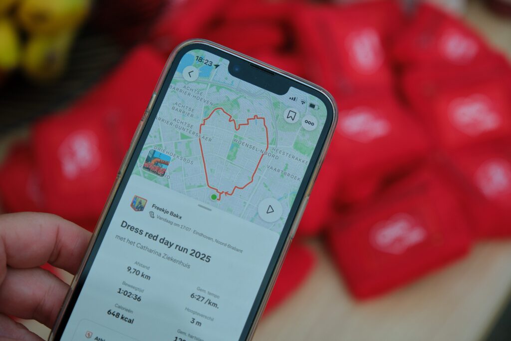 Hand houdt smartphone met hartvormige route op hardloop app, Dress red day run 2025. Rode tassen of shirts vaag op achtergrond.