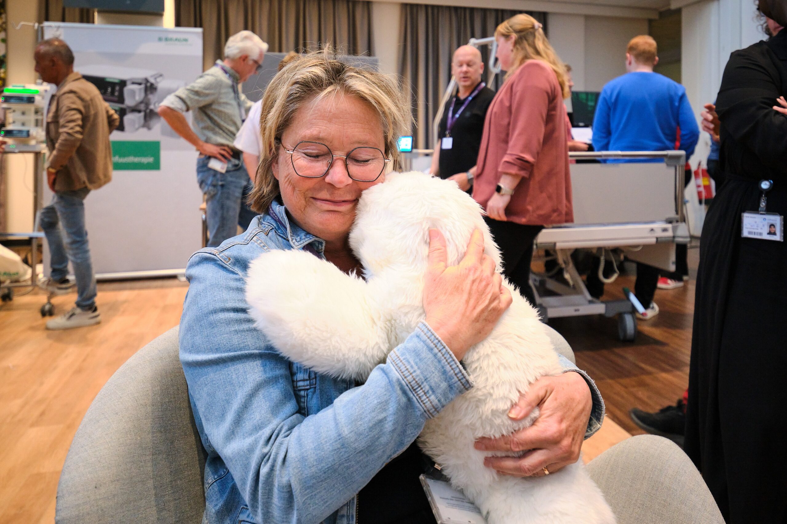 Vrouw met bril knuffelt grote witte robotknuffel in stoel bij Catharina Ziekenhuis. Mensen praten op de achtergrond.