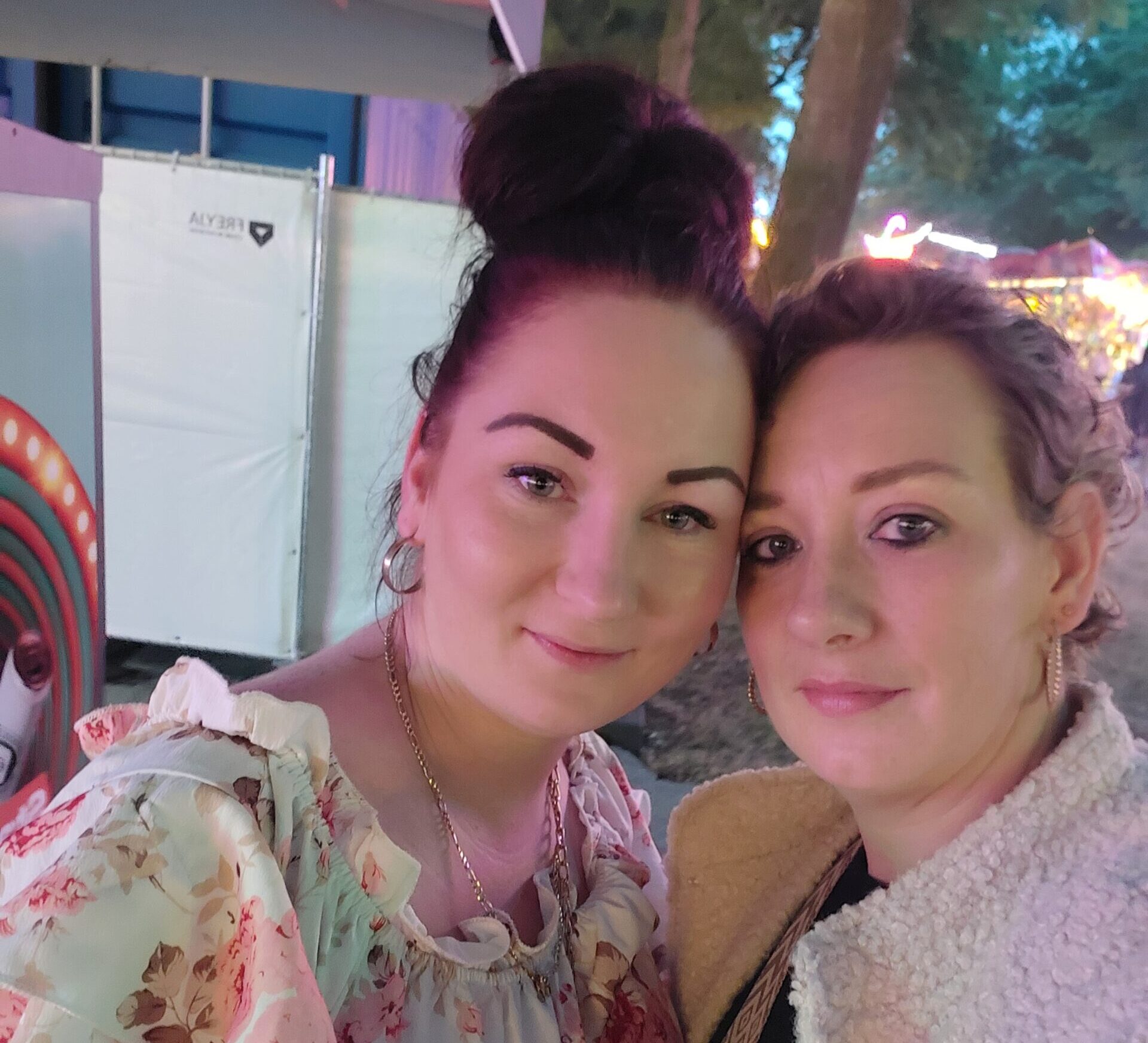 Twee vrouwen maken buiten een selfie. De een draagt een witte bloementop, de ander een lichte krik.