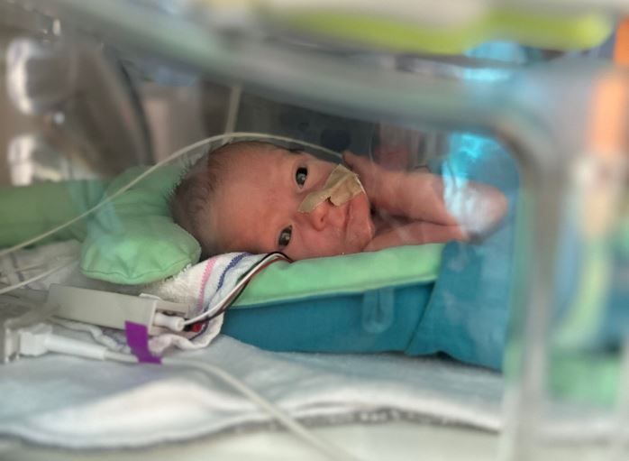 Pasgeboren baby in couveuse met sonde, omringd door medische apparatuur en zacht beddengoed, handje omhoog.