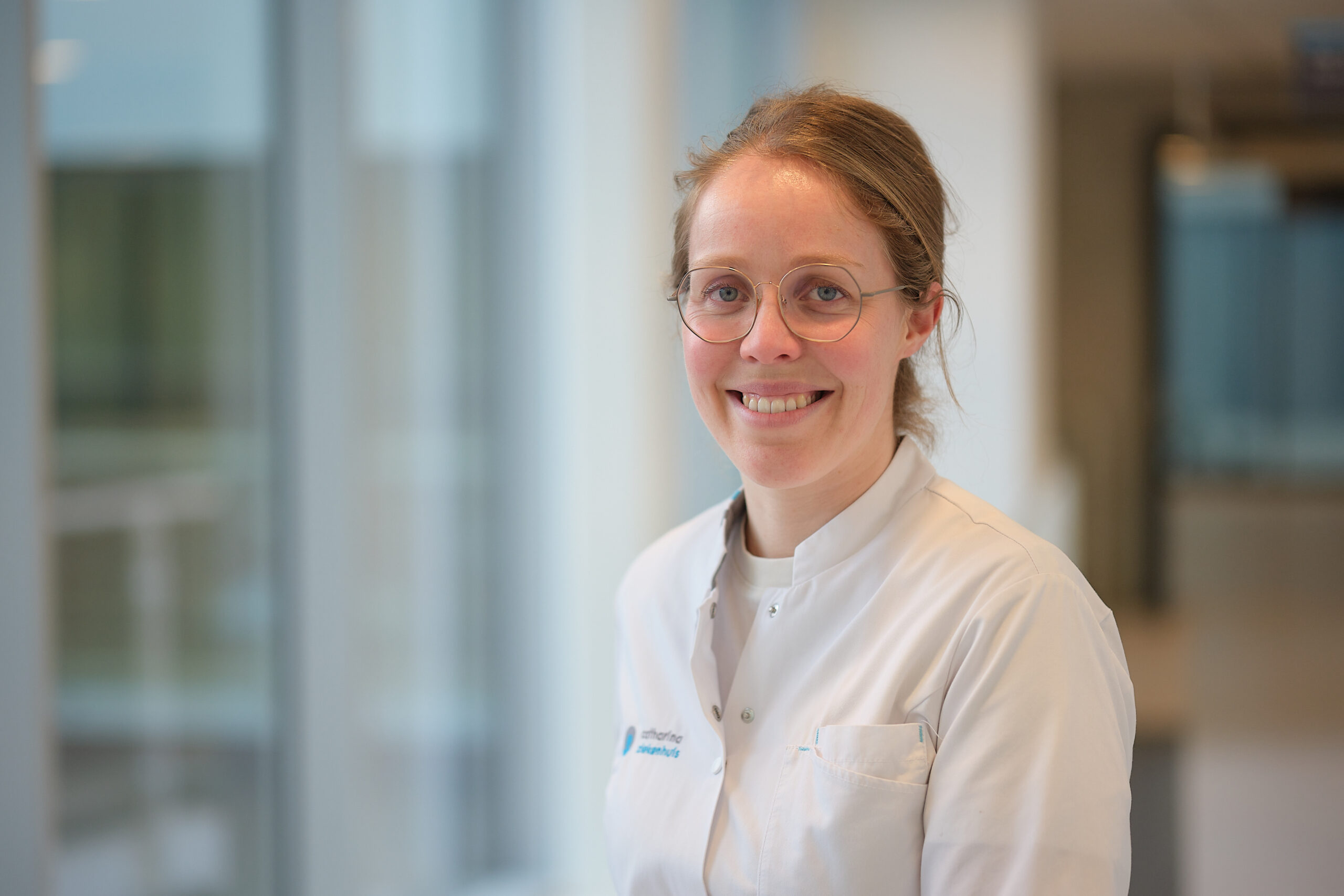 Vrouw met bril en witte jas lacht in lichte bende met grote ramen. Mogelijk bespreekt ze chemotherapie bij darmkanker.